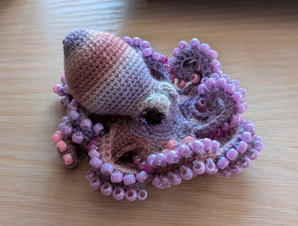 A crochet octopus with pink and purple stripes on the body, and pink and purple beads ss suckers.