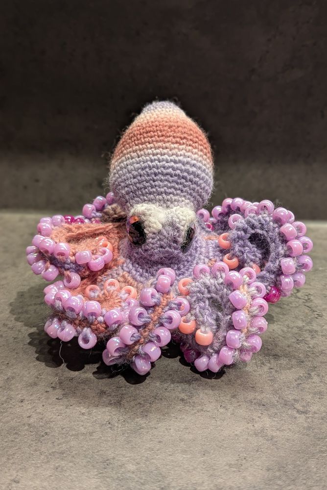 A crochet octopus, with pink and purple striped body, large purple and pink eyes, and purple and pink beads for suckers.