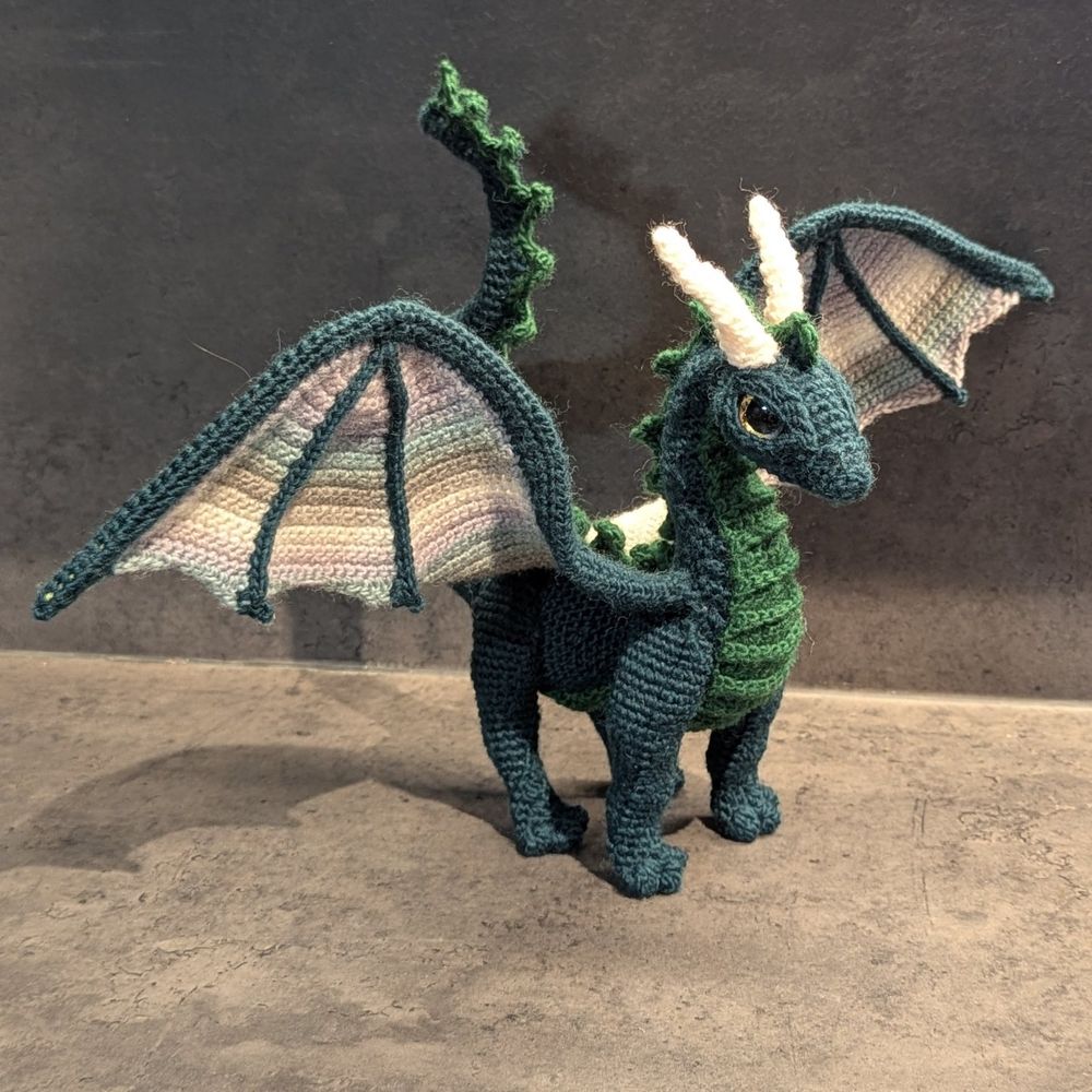A crochet dragon. He's dark green with brighter green belly and back spikes. He's got wings in stripes of grey and greens, four legs, white horns and golden eyes.