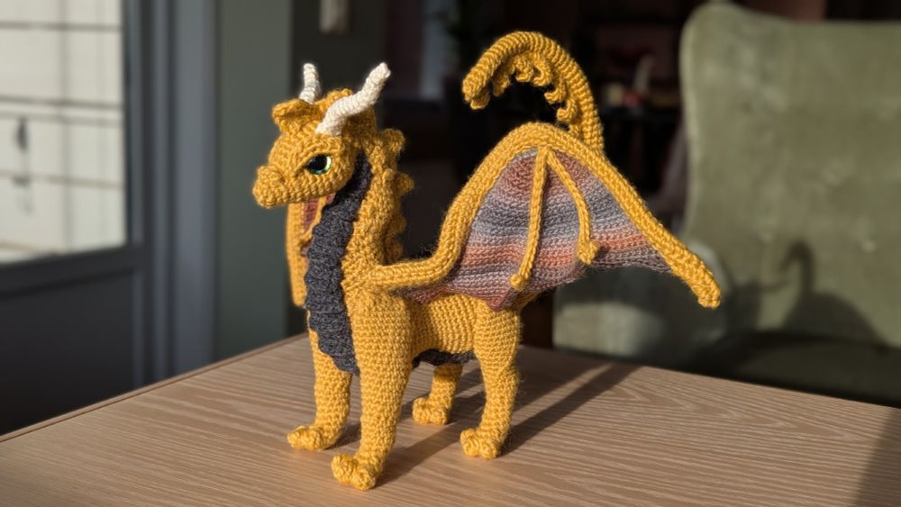 A yellow crocheted dragon, with white horns, dark grey belly, and grey and brown striped wings. 
