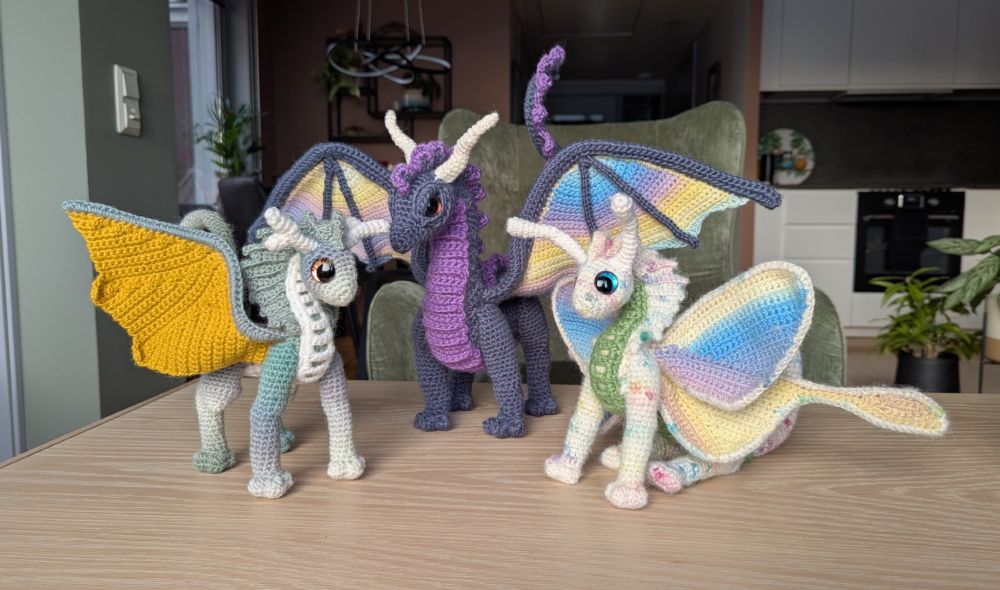 Three crocheted dragons posed together. One has a green-and-white striped body, golden wings, white belly and white horns. The next is larger than the other two, has a blue body, purple belly, rainbow pastel colored wings and white horns. The last one is the only one who sits on his bum, he has a white body with pops of pastel color, pastel rainbow butteryfly-style wings, green belly, and white antennae instead of horns. 
