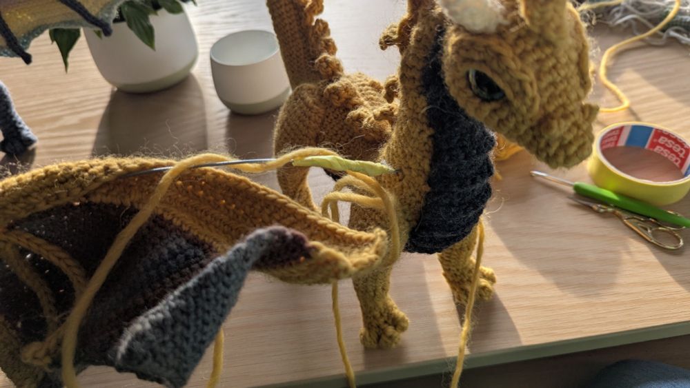 A yellow crochet dragon being stitched together, a bit of wire pokes out of a shoulder.