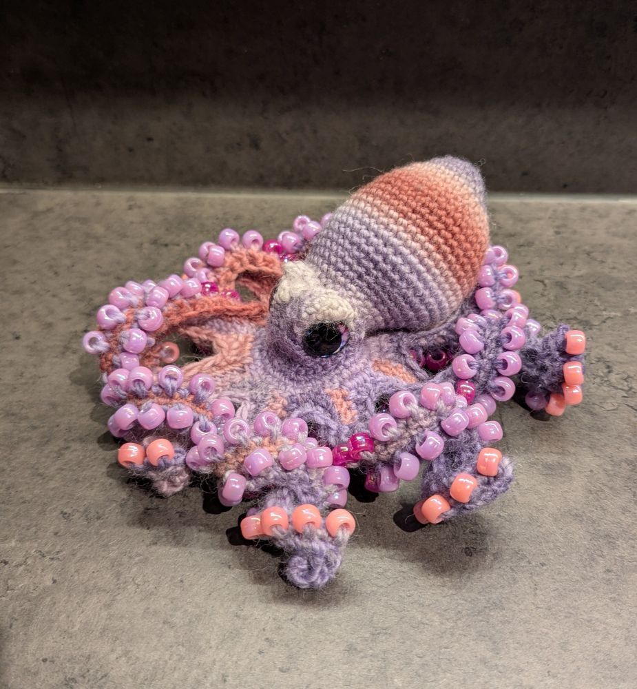 A crochet octopus, with pink and purple striped body, large purple and pink eyes, and purple and pink beads for suckers.
