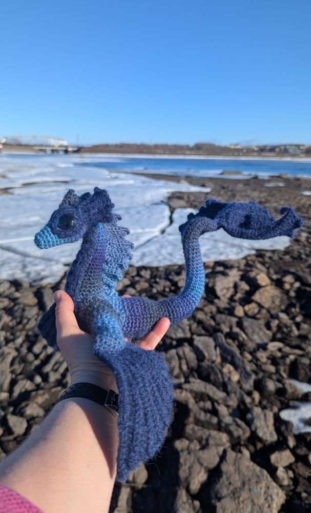 A hand is holding a blue crocheted sea dragon (a dragon-y body with large flippers instead of wings)