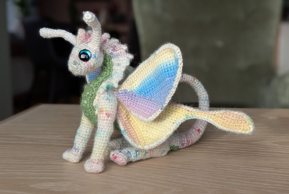 A crocheted dragon sits on his bum. He has a white body with spots of pastel colors, pastel striped butterfly wings, green belly and white antennae. 
