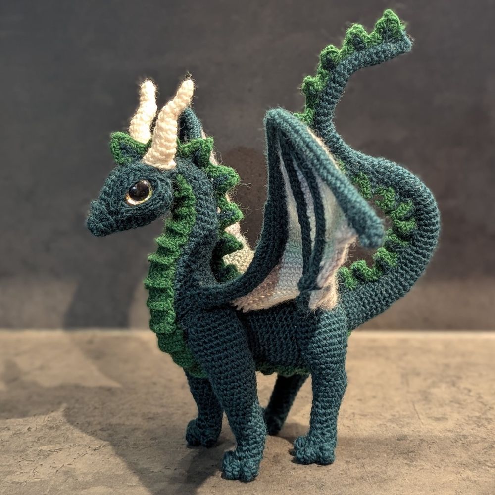 A crochet dragon. He's dark green with brighter green belly and back spikes. He's got wings in stripes of grey and greens, four legs, white horns and golden eyes.