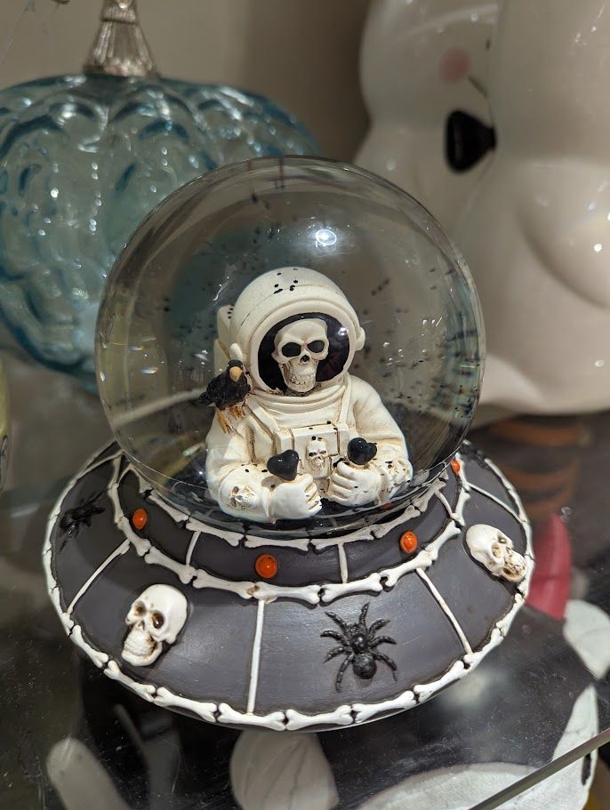 A snow globe shaped like a UFO. There are bones, spiders and skulls around the edge of the UFO. In the globe is a skeleton astronaut. It has a crow on its shoulder.
