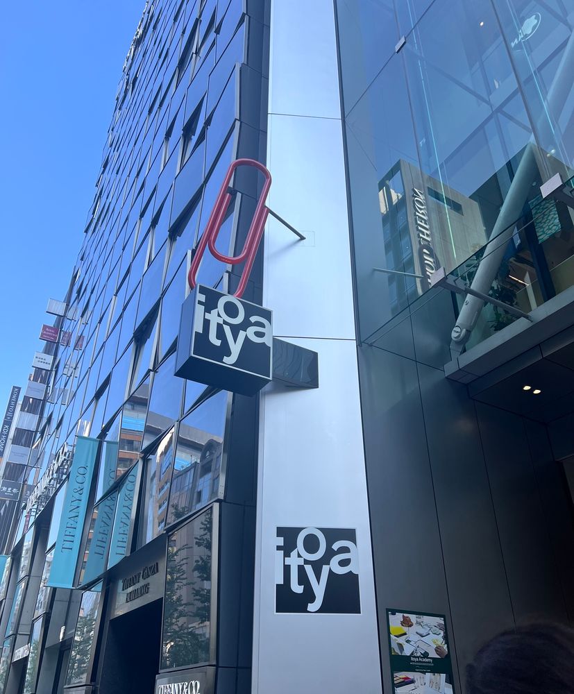 A sign sticking out of a block of very tall glass walled buildings. It’s for a store called itoya, which is a stationery store. Above the shop sign is a statue (?) of a red paper clip. 