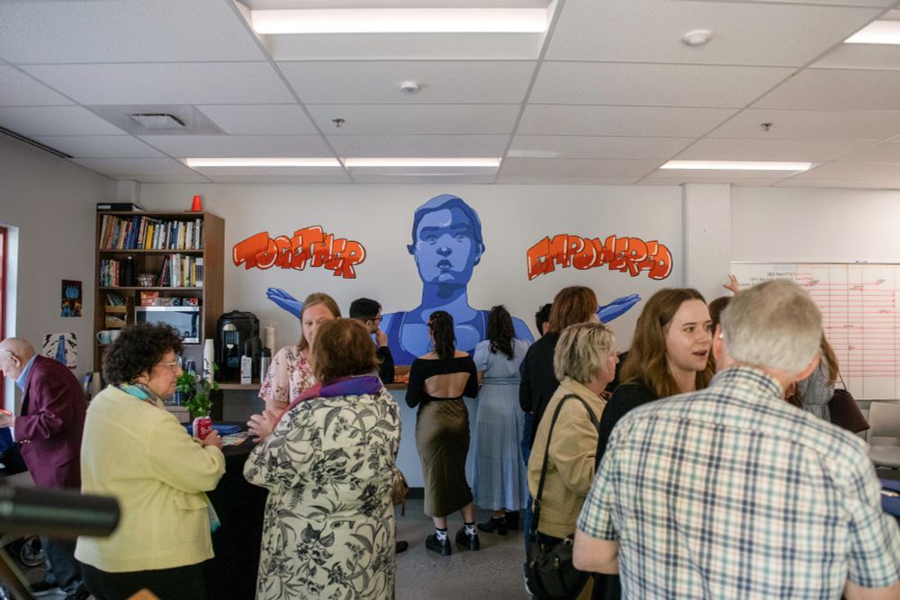 Photo of the crowd at the event, centered on the mural on CHASI's wall. The mural depicts a figure, painted in blue, holding up their hands like scales. Above each hand is a word, saying "together empowered."