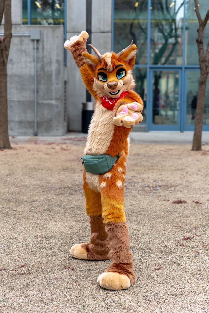 The image shows a person wearing a brightly colored fox-deer fursuit, standing outdoors in front of a modern building with large glass windows. The fursuit has orange and brown fur with lighter cream-colored patches, large green eyes, and visible teeth. The character has antlers, a red bandana, and a green waist pouch. The person is posing energetically with arms extended. The background features trees with bare branches and a gravel surface.