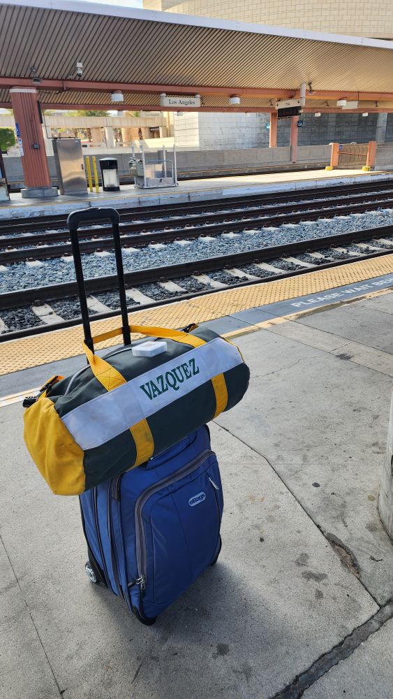 A small rolling blue suitcase with a green and yellow duffel bag sitting on a train platform outside in LA. THE NANE Vazquez is embroidered on the duffel bag. 