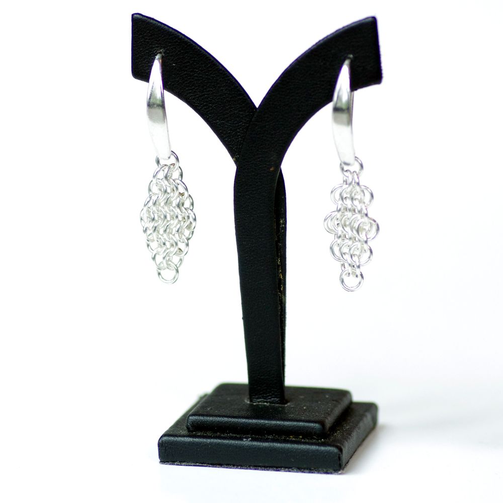 A pair of sterling silver chainmail earrings on a Y-shaped black display stand. Each earring has a thick hook above a diamond-shaped mesh of woven maille.