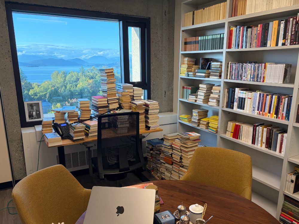 Picture of office with two desks, one covered with piles of books and a bookshelf in the background. 