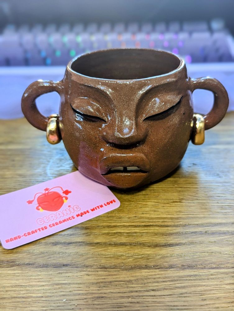 A close-up image of a hand-crafted ceramic planter in the shape of a face with a serene expression, featuring closed eyes, a slightly open mouth, and prominent ears adorned with gold earrings. The planter has a textured, earthy brown surface with a glossy finish. A pink business card with an illustration of a face and the text "Ceramic: Hand-Crafted Ceramics Made with Love" is placed in front of the planter on a wooden surface. The background is softly blurred, showing hints of a keyboard with rainbow-lit keys.