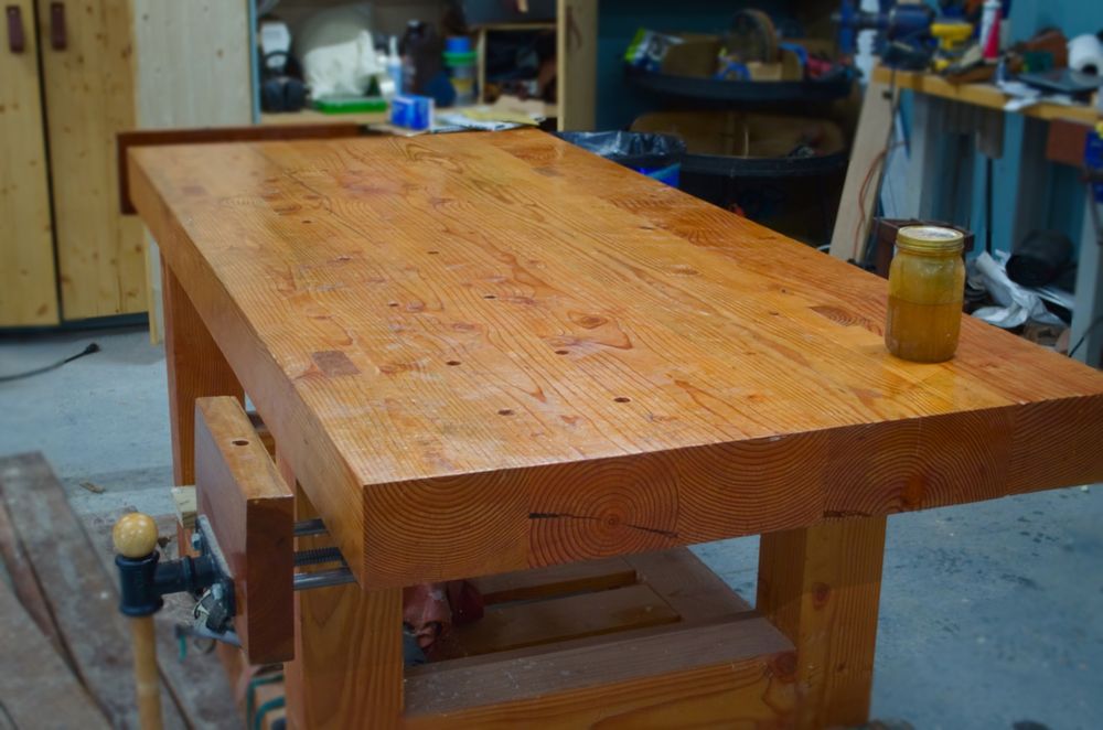 A large, warm amber colored, wooden woodworking table made of fir beams with wood clamps attached to two corners that has had its top freshly reoiled.