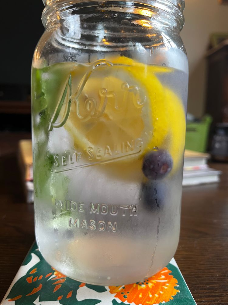 A large jar filled with ice water, lemon slices, frozen blueberries, and fresh mint.