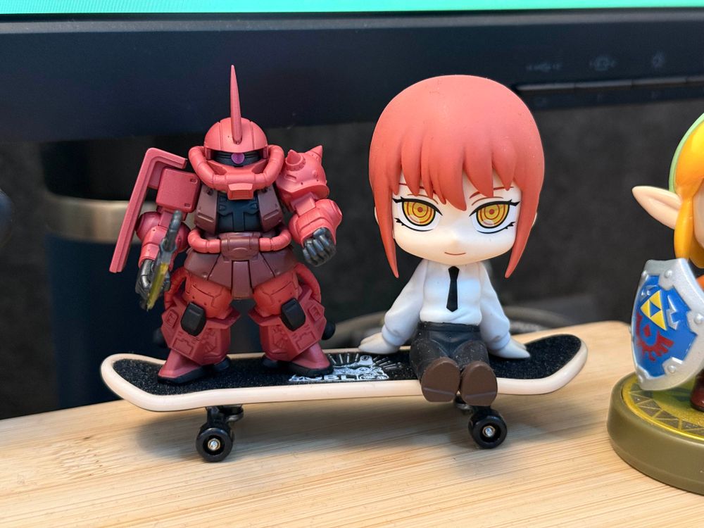 Miniature figures of Char’s Zaku from Gundam and Makima from Chainsaw Man sitting on a Studio Ghibli finger skateboard.
