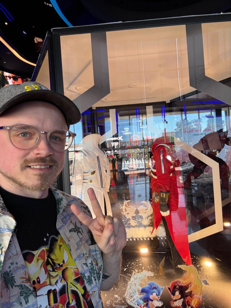Me at the Irvine Spectrum theater standing in front of a display case holding Jim Carrey’s costumes from Sonic the Hedgehog 3. I’m wearing a t-shirt with Super Sonic and Super Shadow standing outside a combination Pizza Hut/Taco Bell.
