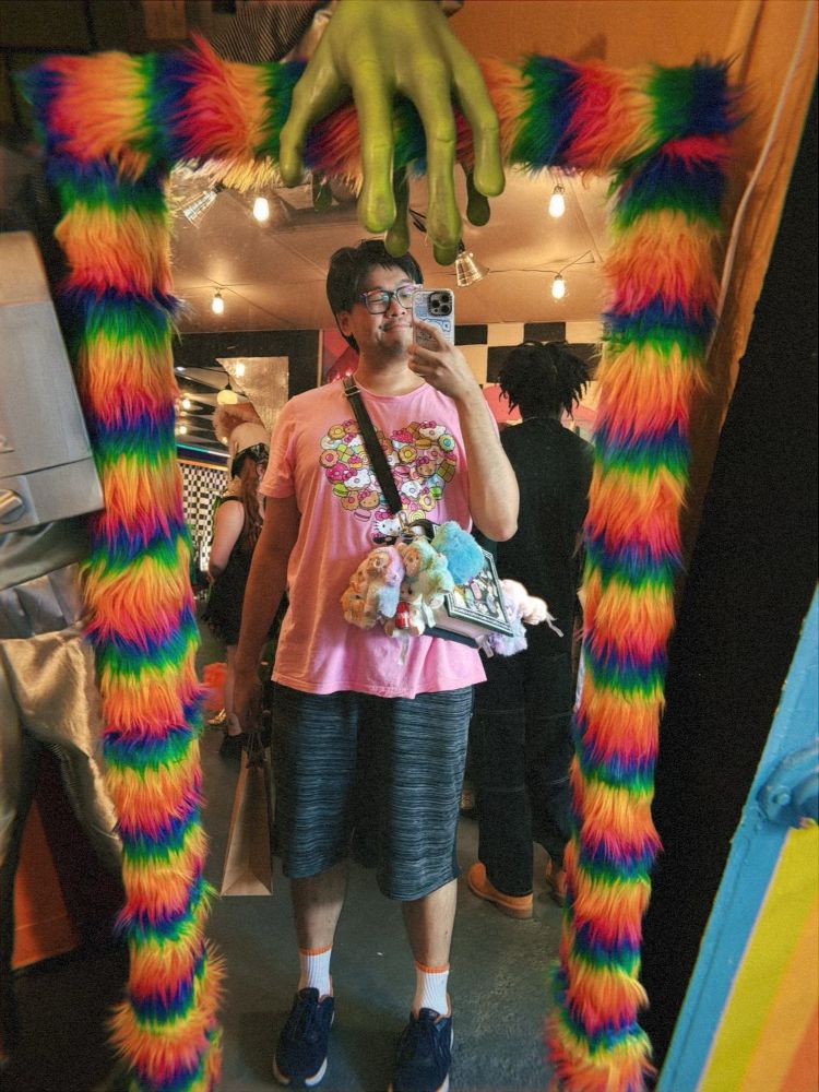 A mirror selfie that sora took at She Comes In Peace. The mirror has a fuzzy rainbow border and a green alien hand on top. sora is wearing a cute pink Hello Kitty t-shirt and an ita purse. On the purse are multiple keychains, including Labubus, Skullpandas, and Miniso Disney plushies.
