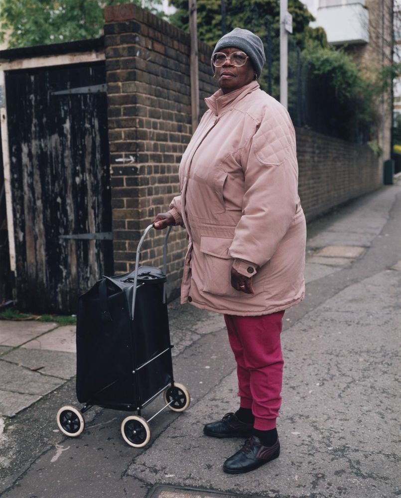 An older woman stands at the entrance to an alley. She is wearing a pink coat and glasses, and she is posing with a black shopping trolley.
