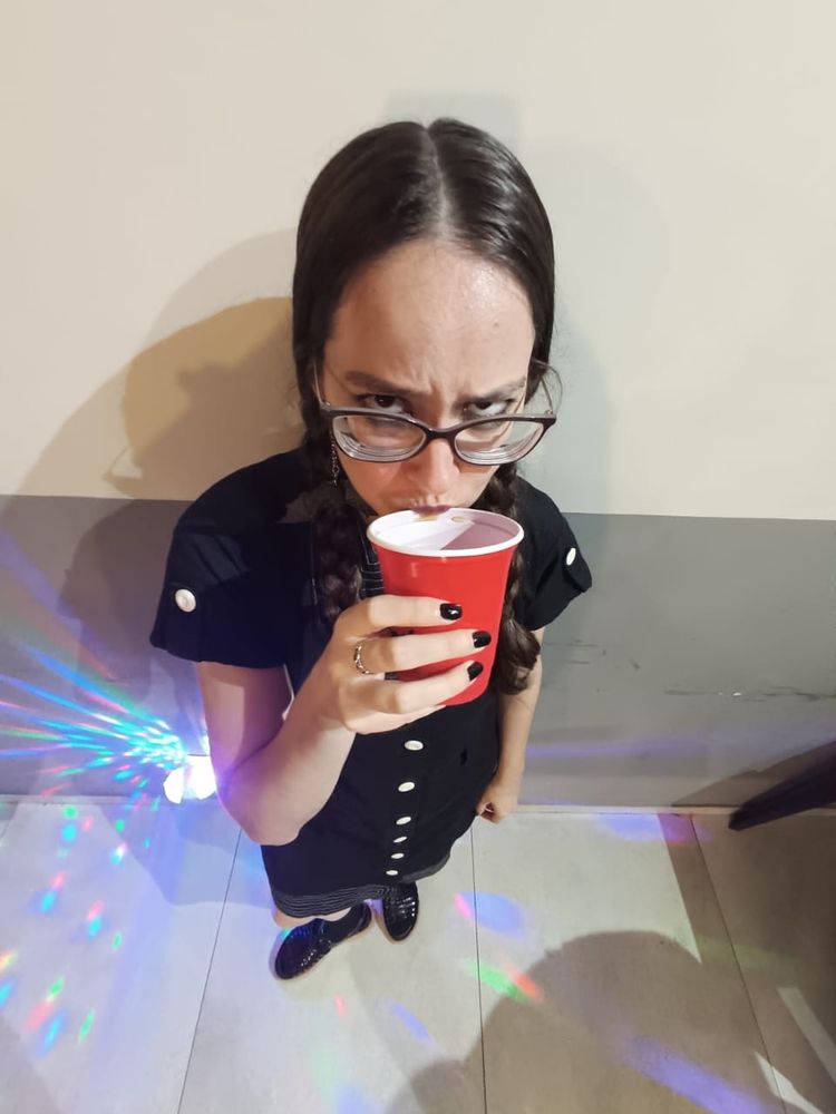 Foto tirada de cima no zoom 0,5, ou seja, corpinho e cabeção. Roupa igual da priemira foto. Tô de sapato preto e meia preta, segurando um copo vermelho, que ta encostado na minha boca. Tô de óculos e com uma expressão esquisitíssima.