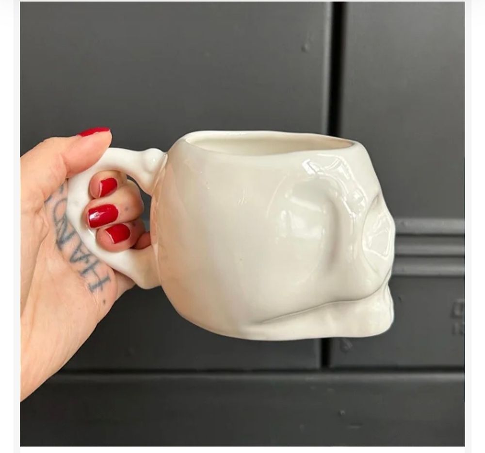 Idem porém de lado dando pra ver a parte de trás da caneca que tem uma alça, que está sendo segurada por uma mão branca, com tatuagem na palma e unhas pintadas de vermelho.
