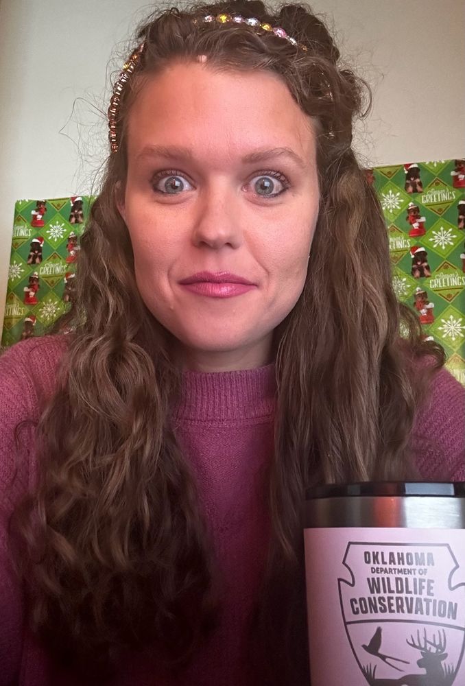 I  am a white woman with curly brown hair and makeup on with a pink sweater and am sitting in front of Christmas paper with my OK Dept. Of wildlife mug. 