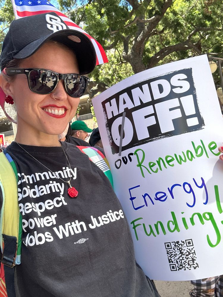 This image shows a white woman with a T-shirt that says community solidarity respect, power jobs with justice. She’s holding a sign that says hands off our renewable energy funding. She’s wearing a San Diego Padres hat with the roses on it.