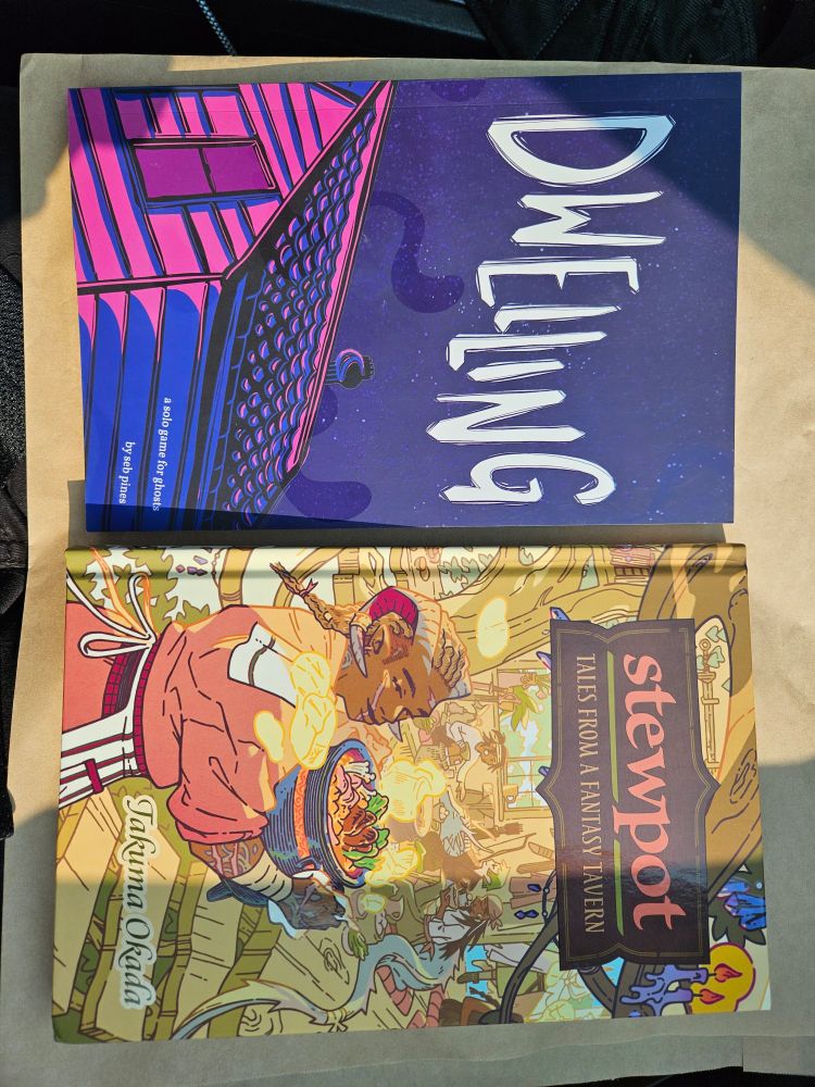 Side by side spread of two tabletop role-playing game booklets on a brown paper background. The book on the left (or top) is a blue, blue, and pink design of a house with the title "Dwelling" in white. The book on the right (or bottom) has the title "Stewpot: tales from a fantasy tavern" and has a cozy fantasy tavern with wooden accents. The character in the foreground is wearing orangeish clothing and carrying a pot of stew.