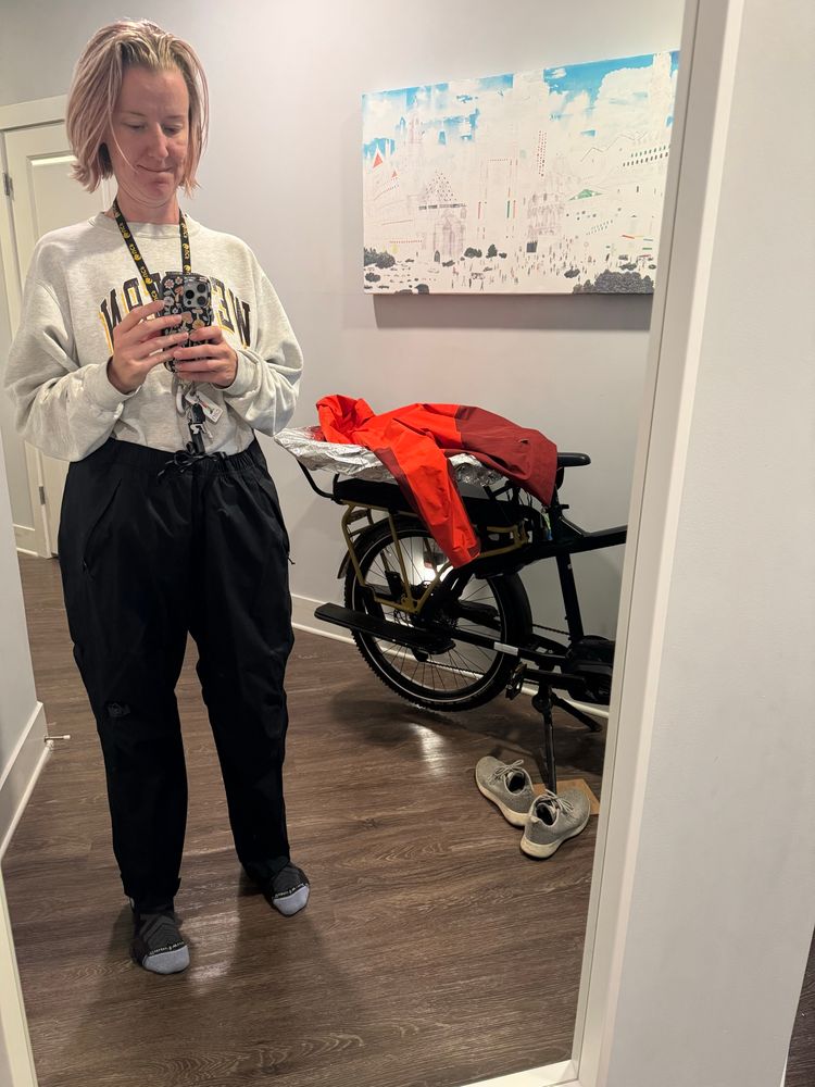 Photo of me wearing rain pants over my pants and my cargo bike with a rain cover to protect the rear seat so my 1st grader doesn’t have to sit on a wet seat when I pick him up from school.