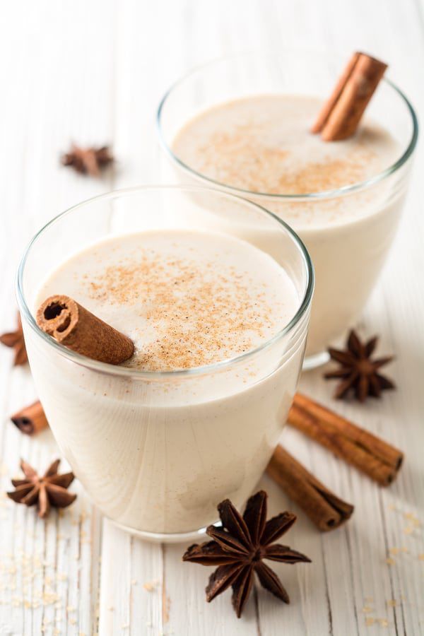 Two glasses of eggnog with star anise and cinnamon surrounding them on a white wooden background