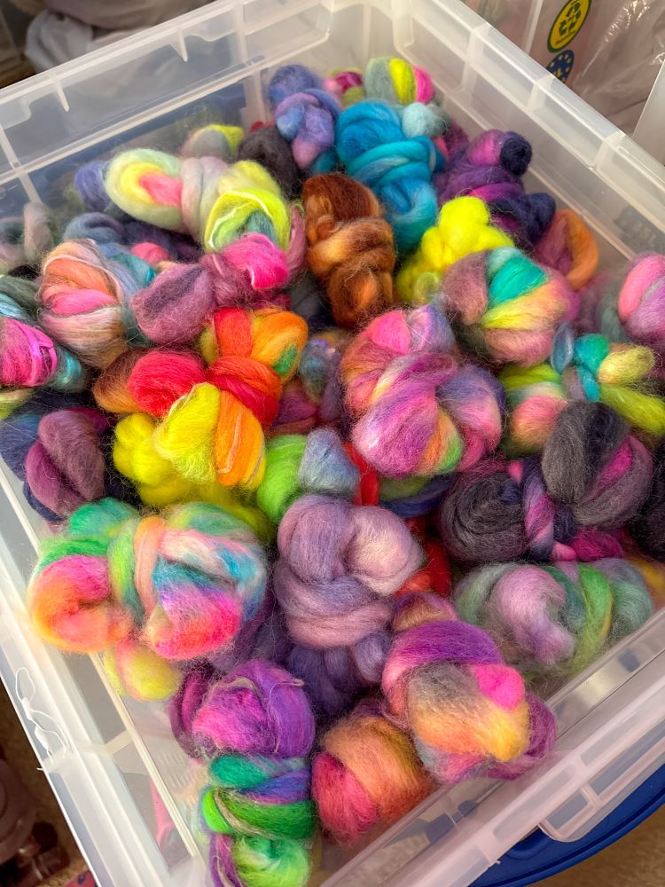 A tub full of little strips of hand dyed fibre braids, rolled up into little nests. There are too many colours to mention 🌈 