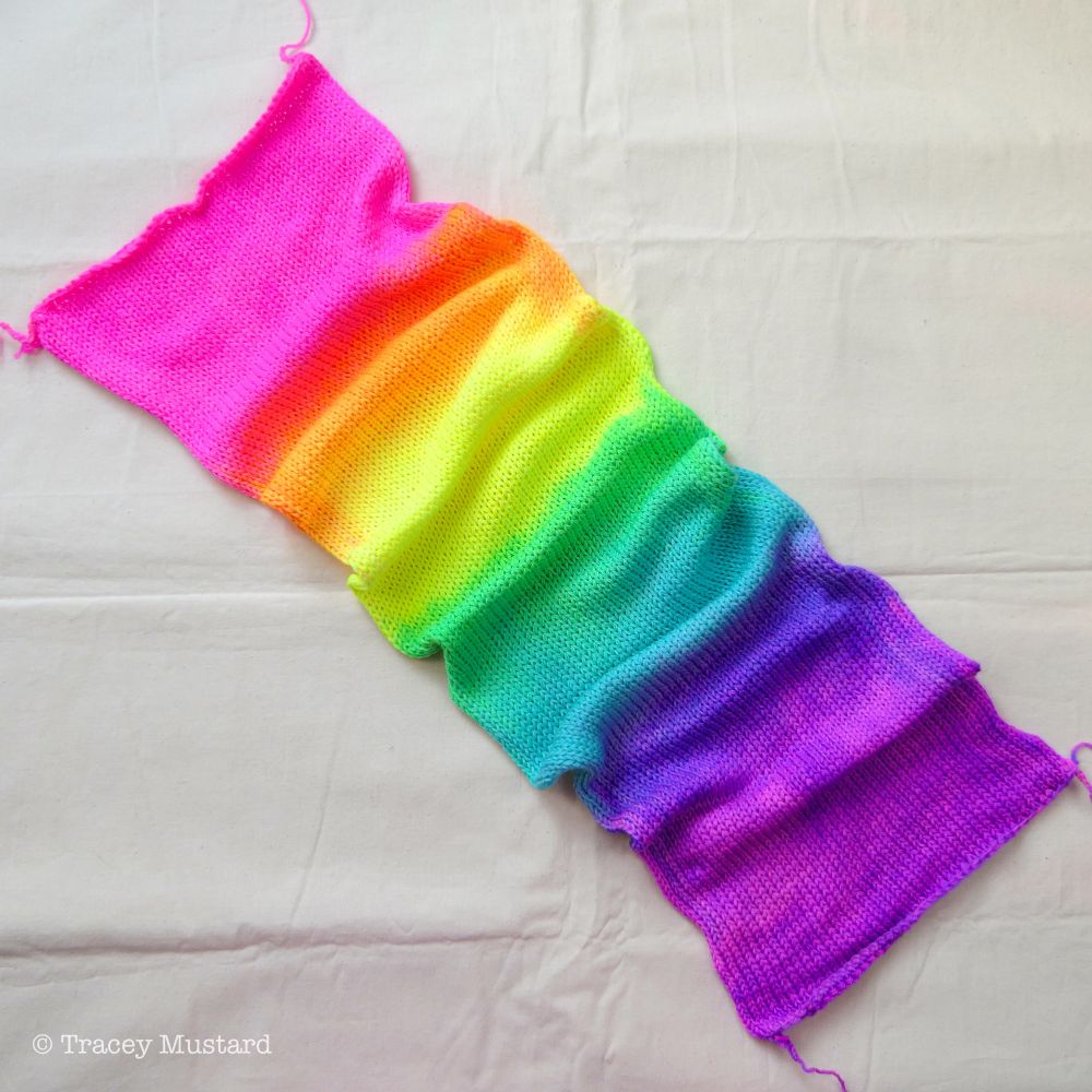 A top-down of a hand dyed sock blank. It’s been hand-painted in a neon rainbow gradient moving through the blank from top to bottom, from (all neon dyes) Pink, into Orange, Yellow, Green, Aqua Blue, Lavender, and ending with Purple.