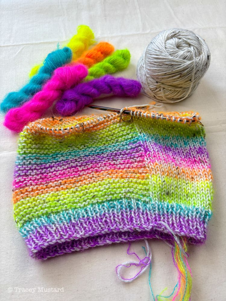 In the top left corner is a little pile of bright Mohair micro-skeins in neon pink, orange, yellow, green, blue, and purple. In the top right is a cake of undyed tweedy DK yarn, and in the middle is a glorious knitted hat WIP on circular needles. A rainbow moves through the hat in stripes as I'm doing four rounds of each Mohair colour, the undyed yarn creating a marled fabric. There are little flashes of black, brown and grey now and then where the nepps are saying hi.