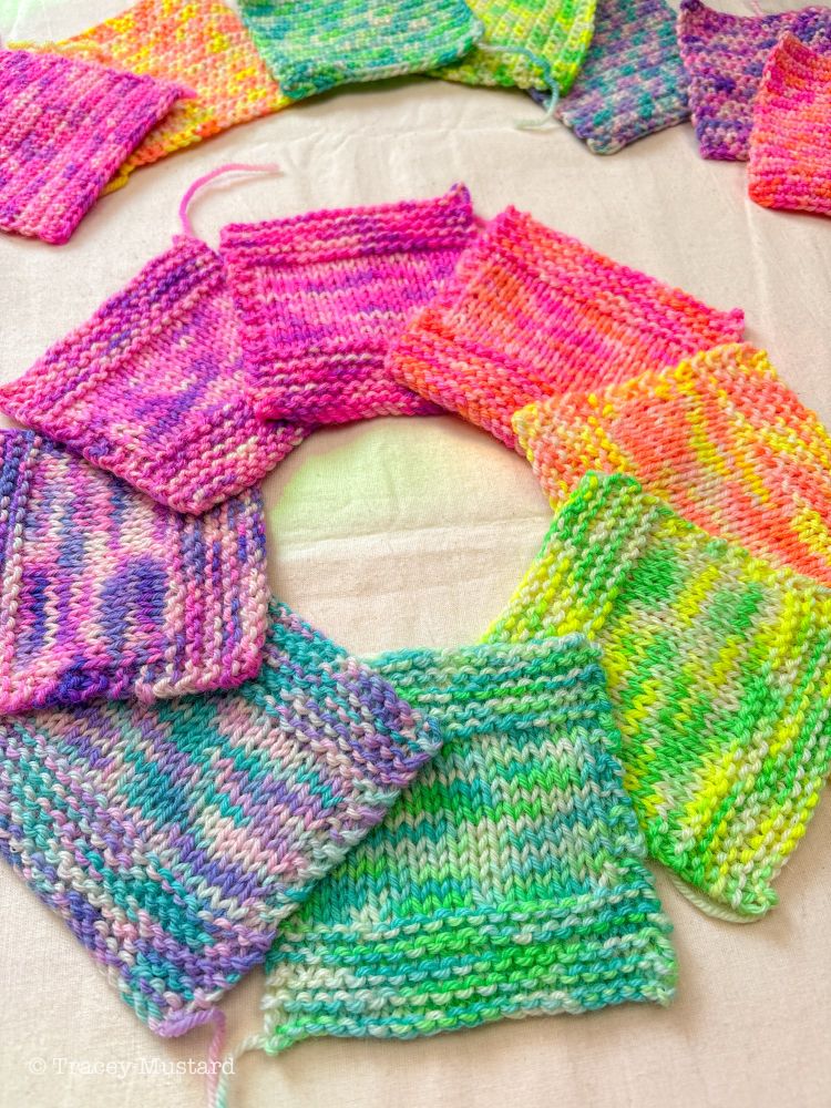 Eight square knitted swatches sit in a circle overlapping one another. The yarn is variegated, with one colourway containing neon pink and coral, then the next containing neon coral and yellow, the next containing yellow and green, continuing like that through aqua blue, lavender, purple and magenta. There are a few crochet swatches peeking out at the top of the picture. 