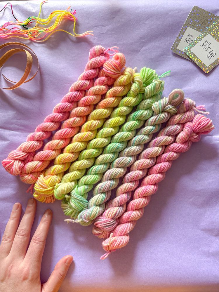 Eight twisted skeins of variegated hand dyed yarn sit side by side diagonally; a neon pastel rainbow runs through the colourways from top to bottom. My left hand is resting in the bottom left of the scene to remind everyone that I'm a real-life person running this small biz, some What Mustard Made yarn labels are in the top right, and some kraft paper ribbon and precut pieces of handdyed yarn are in the top left. Everything is sitting on pastel lilac tissue paper which I use to wrap up orders.