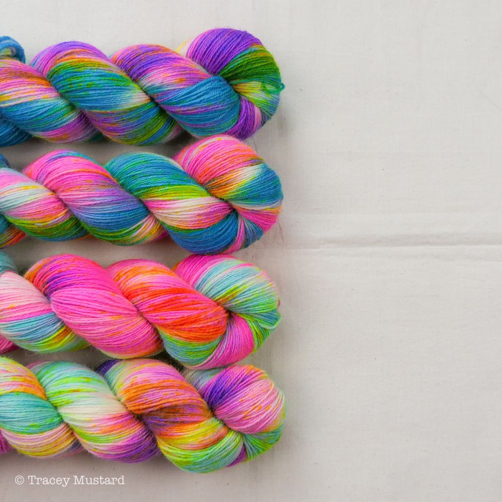 Four twisted skeins of hand dyed yarn sit to the left of the photo, with half of them cut off. The yarn is dyed in a variegated manner, using neon purple, turquoise and neon yellow speckles in Bliss (top one), neon pink, turquoise and neon yellow speckles in Euphoria (next one down), neon pink, aqua blue and neon yellow speckles in Joy (next one down), then neon purple, aqua blue and neon yellow speckles in Rapture (the last one). They are sitting on a cream linen cloth, which I haven't ironed because life is far too short and my time is too precious to be spending it ironing. I also find the creases very handy for squaring up my photos! 😅🤷🏻