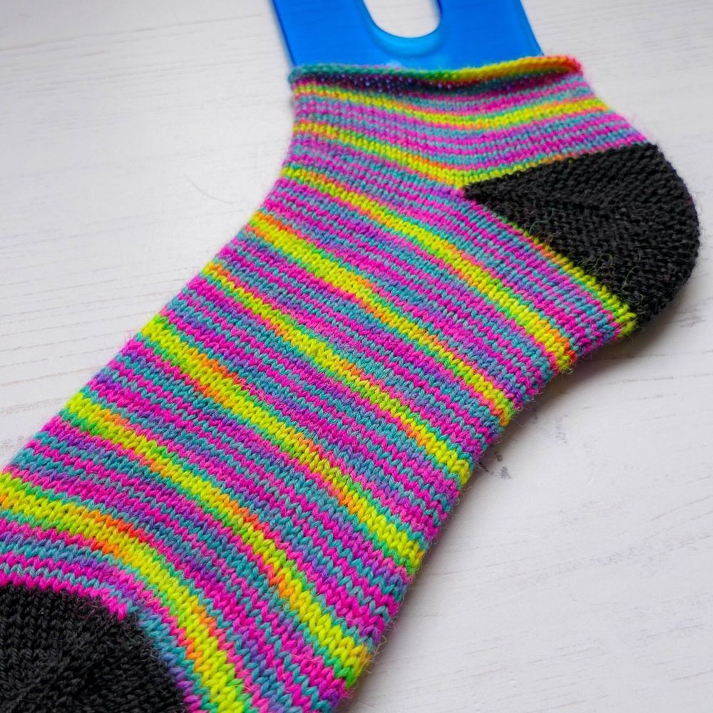 The most beautiful hand knit sock posed on a sock blocker - the way the colourway is dyed creates microstripes of neon pink and blue, with slightly thicker stripes of neon yellow with tiny pieces of orange and green at either end of the yellow. The toe and heel are black. 