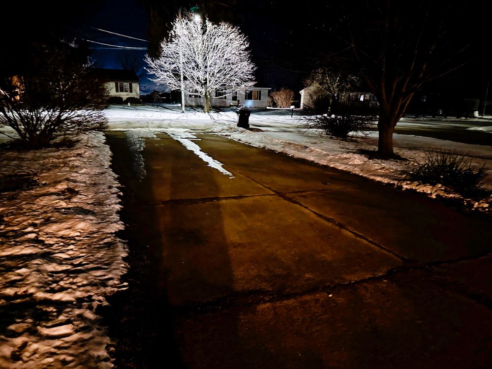 A photo of a driveway, after dark. A tree across the road is illuminated by a street lamp, as is some of the rest of the image. It looks icy.