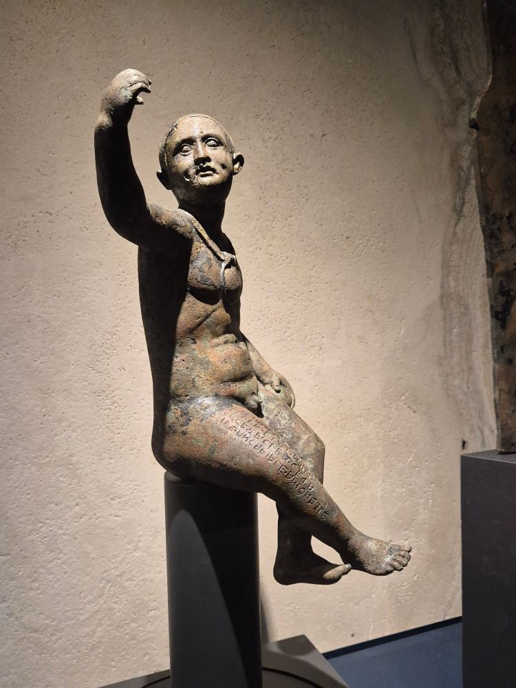 Bronze votive statuette of a sitting chubby baby boy, with a large bulla around his neck and an Etruscan inscription on his right thigh. 