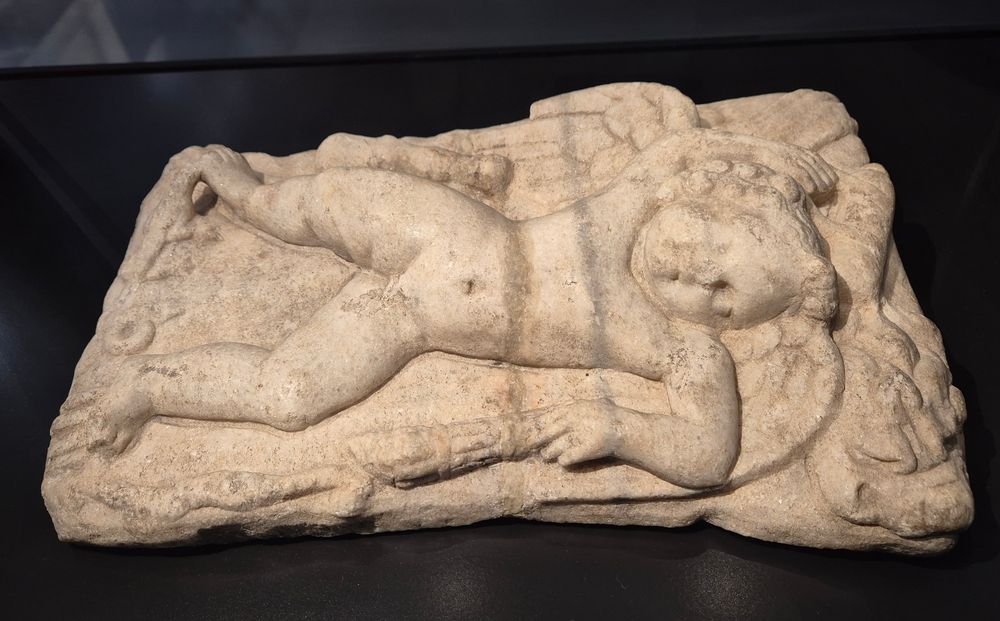 Marble statue on Eros sleeping on a rock. In his left hand, he holds a torch lying next to him. On his right side lies Hercules' club, and the liom skin lies above his head. At his feet, are a snake and a little bird which seems to bite into his right big toe. 