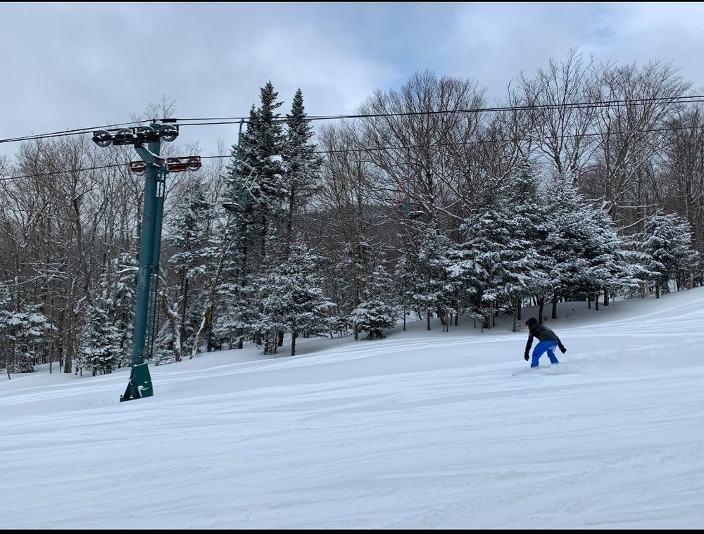 Snowboarding at Bolton Valley Resort on February 13th, 2019. I'm riding down a very snowy trail under a lift, with powder getting kicked up around my board, with no one around (except my friend taking the photo)