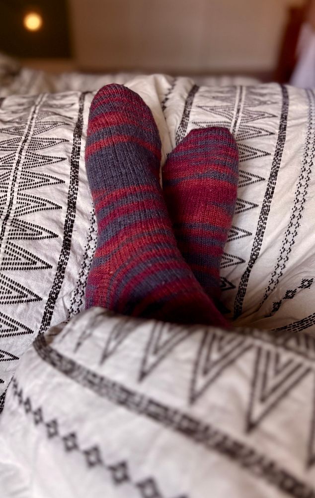 A close up photo of feet in red, orange, and gray wool socks laying on top of a white comforter with black geometric stitching designs. 