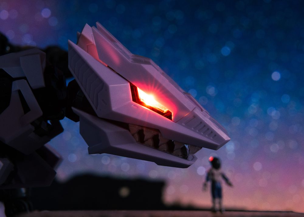 A close-up of a robotic dragon's head with glowing red eyes, set against a starry night sky. In the foreground, a small astronaut figure stands, adding a sense of scale.