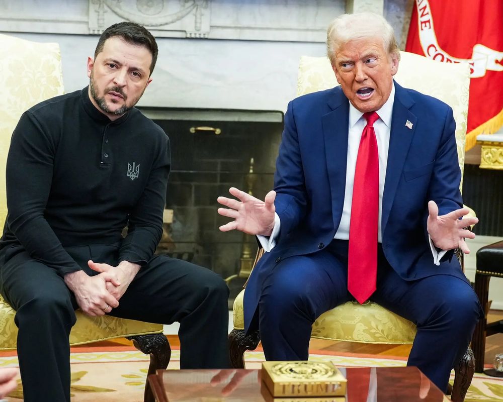 A photograph of Volodomyr Zelenskyy and Donald Trump in the Oval Office. Zelenskyy is wearing all black with the insignia of Ukraine on his shoulder. Donald Trump is wearing a navy suit, white shirt and long red tie. He is red in the face, appearing to shout, and gesticulating. Zelenskyy is staring into the camera, looking tired and exasperated. 

