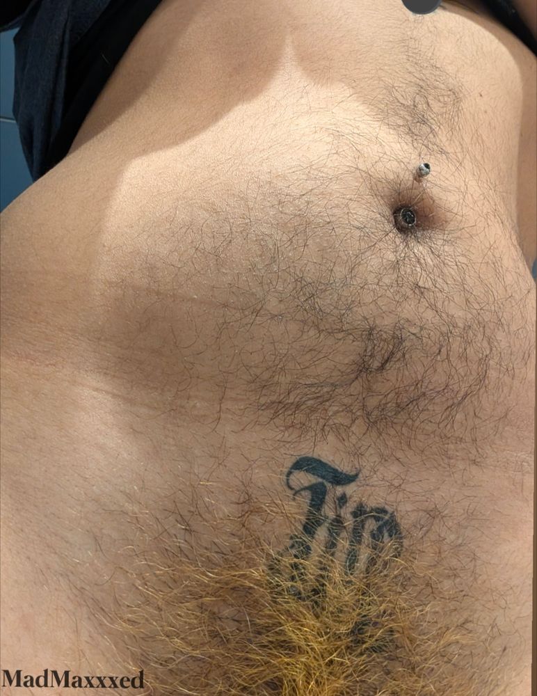 A trans Mexican american faces the camera
In frame is their stomach and the very top of their pubes
They have a black gemstone bellybutton piercing
They have dark brown fuzzy hair on their stomach
Their pubes are orange
They have a tattoo saying FIRE centered above their crotch, below their belly
