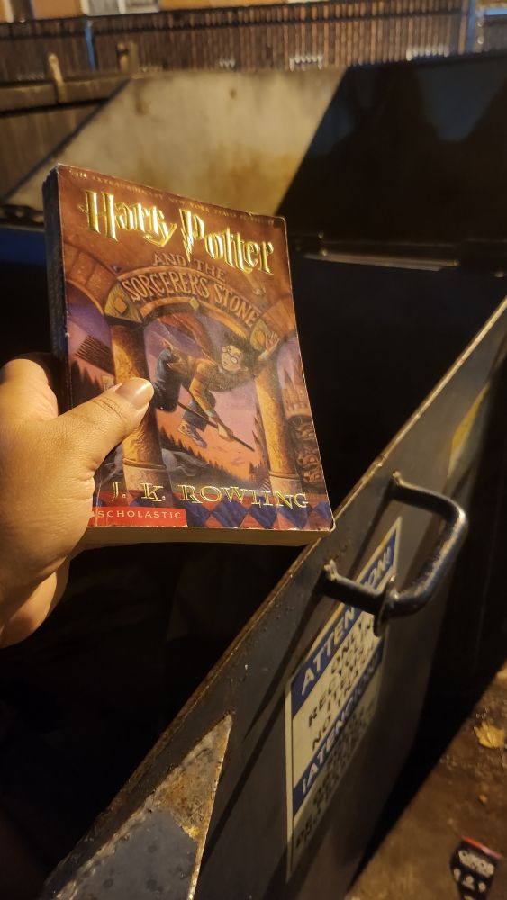 An old worn copy of Harry Potter and the Sorcerer's Stone being held over a dumpster ready to be dropped in. This is a kind act.