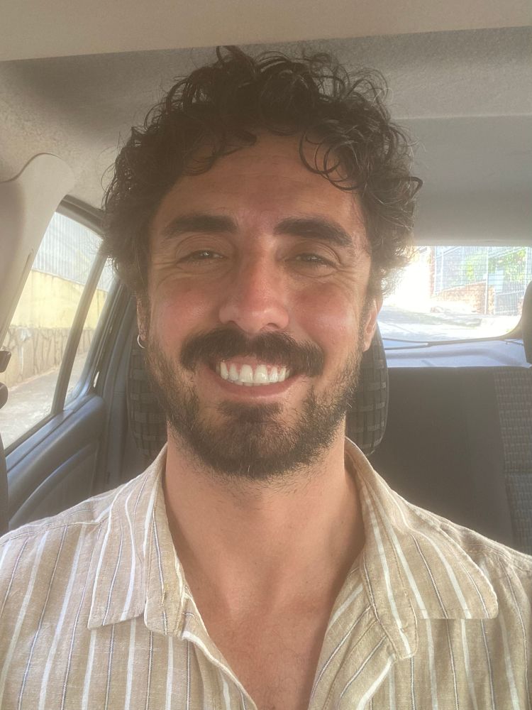 selfie minha tirada dentro do carro. sou um homem branco, de cabelo preto, curto e cacheado, barba e bigode. estou usando uma blusa de linho listrada, em tom pastel e com os botões de cima abertos, e sorrio pra foto