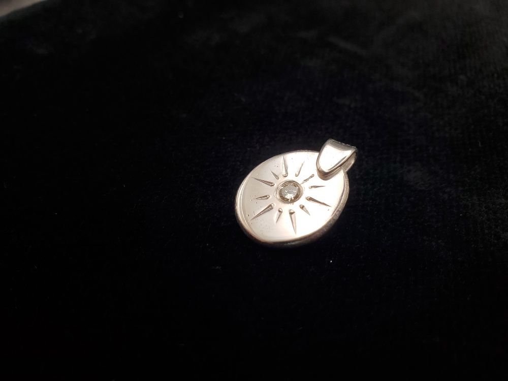 A very shiny silver pendant with a gemstone at its center and an engraved sun pattern surrounding it, on a black background 