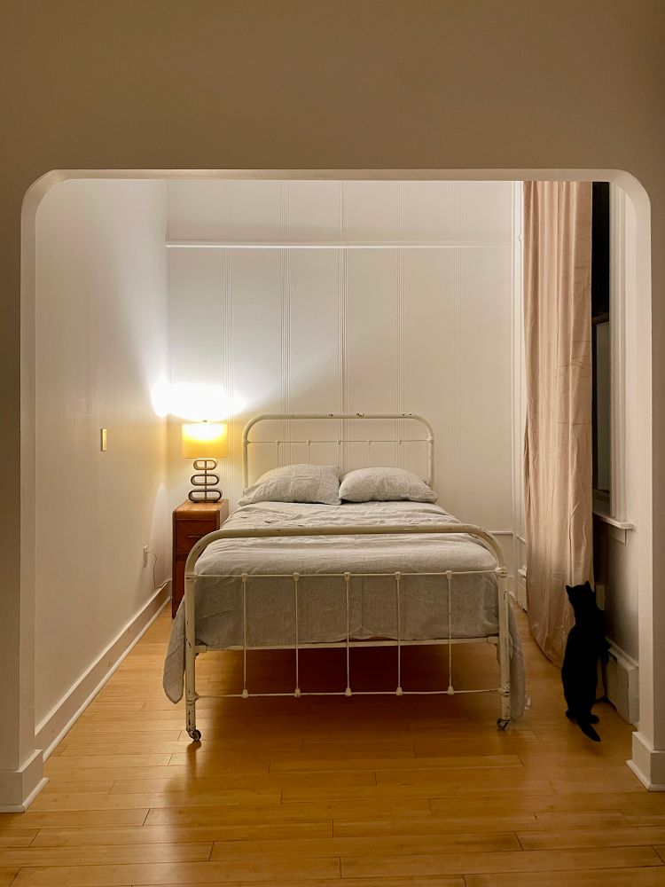 an image of my new bedroom with my white cast iron bed in a cute little alcove. there's a window with a paltry curtain (i'm getting another set to fill it out more!) and dottie my black smoke tabby cat is investigating behind the curtain a bit 